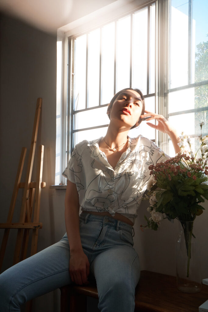 Nadya Inzunza sits by a window bathed in soft sunlight wearing a white blouse with abstract designs and blue jeans. She touches her face and beside her is a vase with fresh flowers.
